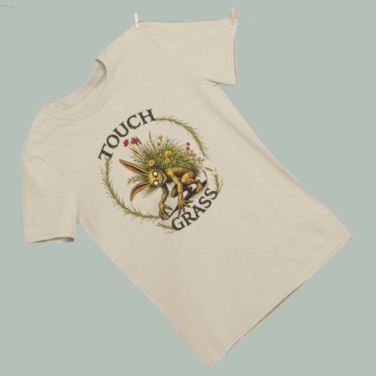 Sand colored heavy unisex tee shirt goblin fairy fae stray sod creature crouching with grass growing from his back the words say Touch Grass. 
